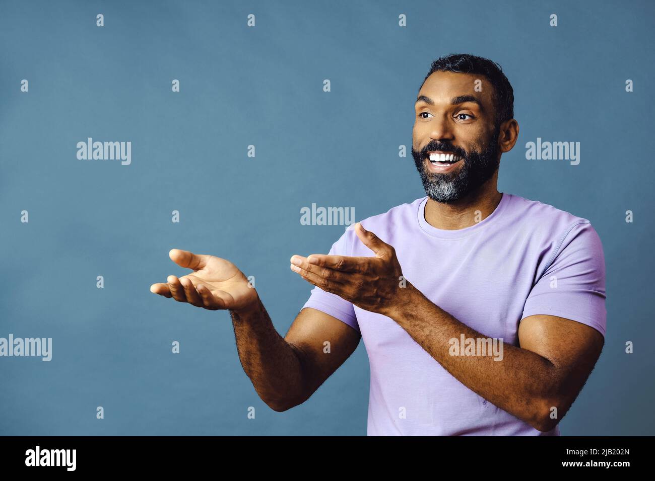 jeune homme noir avec barbe et moustache souriant et regardant loin de l'espace de copie main signal isolé de fond bleu studio Banque D'Images