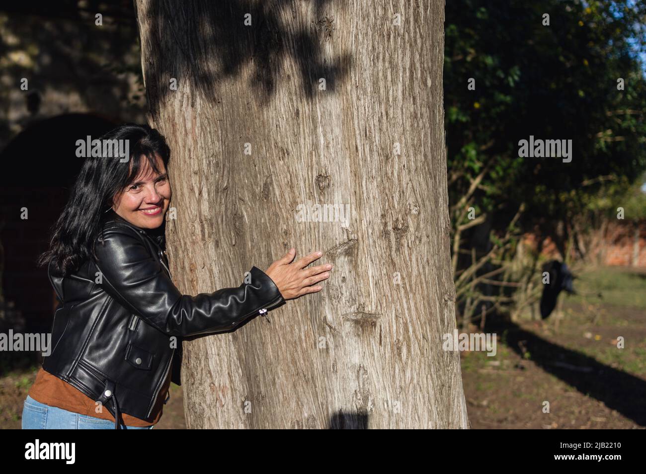 femme adulte de race blanche latine, vêtue d'une veste noire, en plein air regardant l'appareil photo sourire et embrasser un grand arbre, espace de copie. Banque D'Images