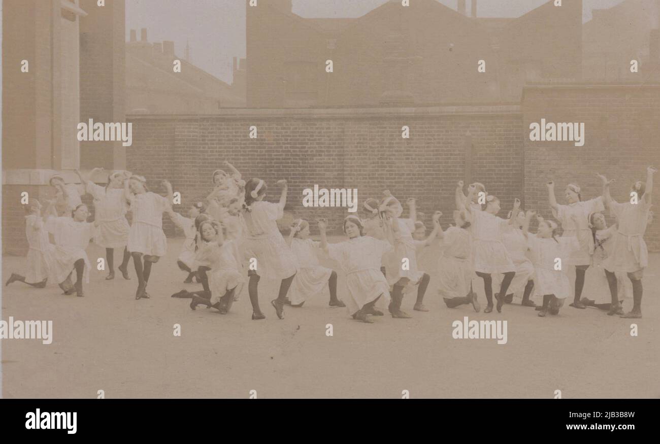 Photo du début du 20th siècle des filles d'école faisant une sorte d'exercice physique ou de danse dans une cour. Toutes ont des robes pâles assorties, des collants foncés et des fleurs dans leurs cheveux Banque D'Images
