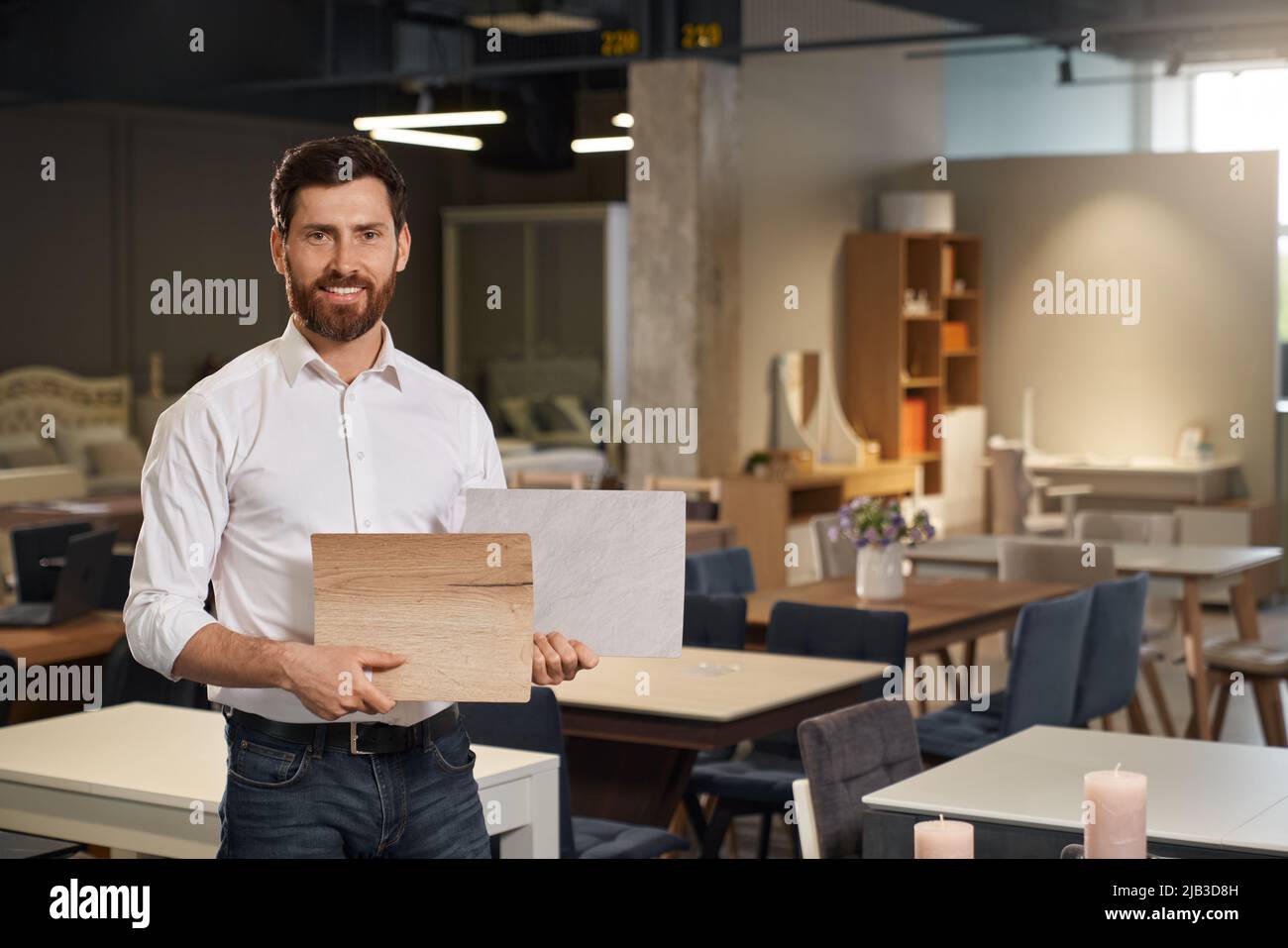 Vue de face de Happy Designer debout, tenant, montrant des échantillons en bois. Beau brunette mâle avec la barbe en train de concevoir, regarder l'appareil photo, sourire. Concept de conception. Banque D'Images Vue de face de Happy Designer debout, tenant, montrant des échantillons en bois. Beau brunette mâle avec la barbe en train de concevoir, regarder l'appareil photo, sourire. Concept de conception. Banque D'Images