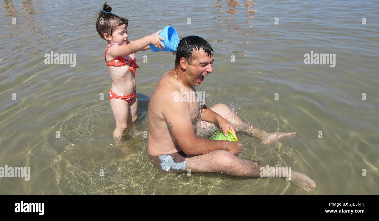 Image d'un père assis dans l'eau criant tandis que sa fille avec un seau jette de l'eau sur son dos. Banque D'Images