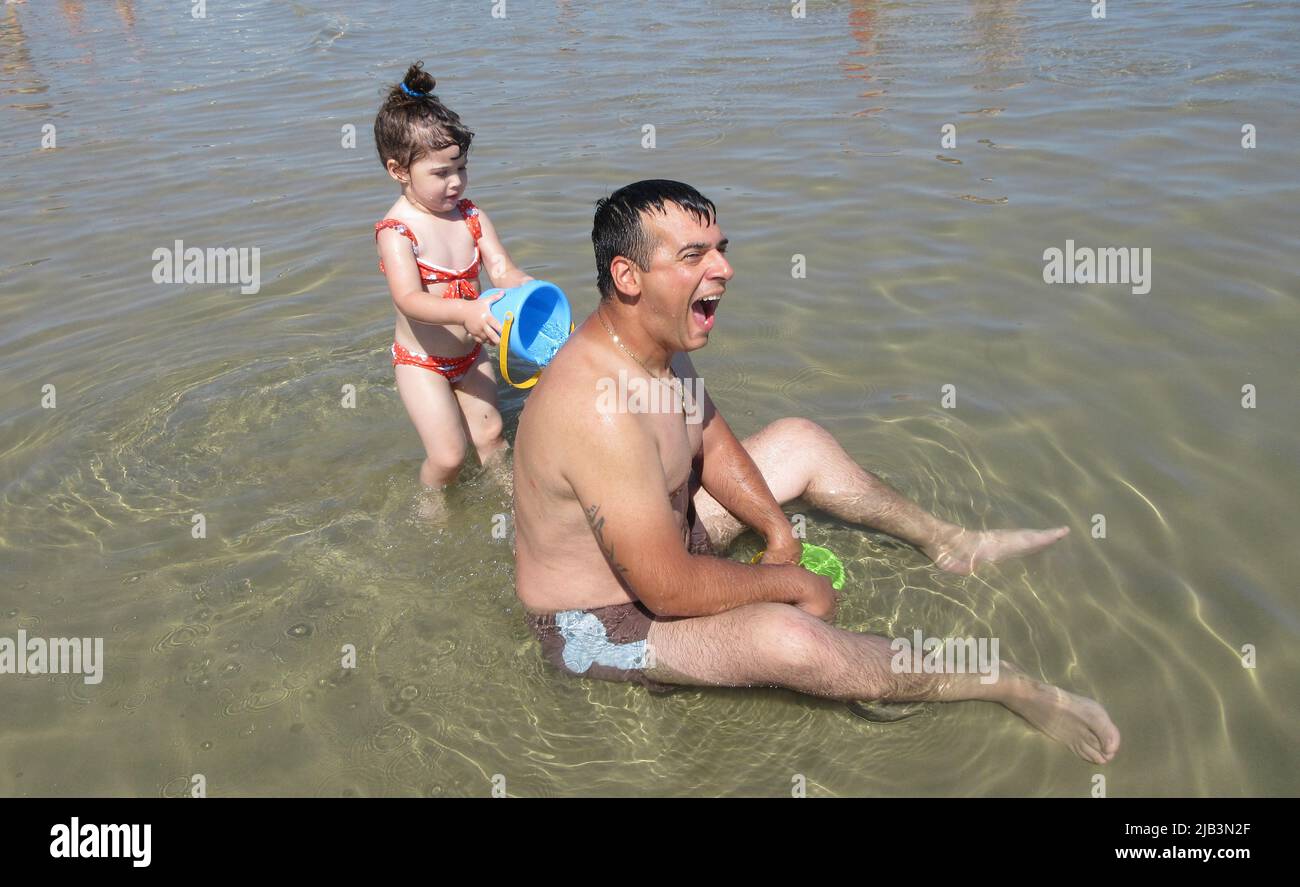 Image d'un père assis dans l'eau criant tandis que sa fille avec un seau jette de l'eau sur son dos. Référence aux températures élevées Banque D'Images