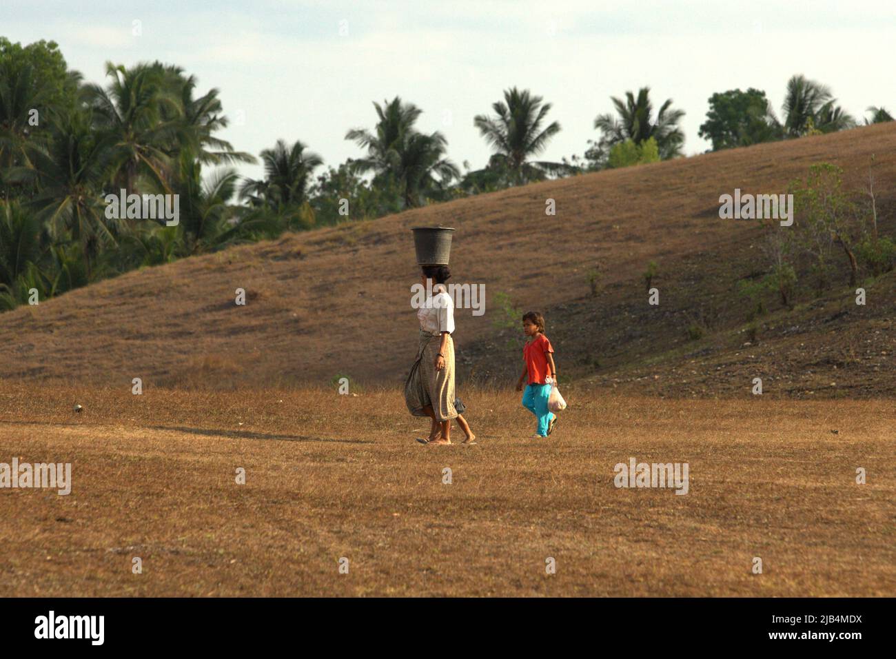 Une femme portant un seau en plastique sur sa tête alors qu'elle marche avec des enfants sur des prairies sèches pendant la saison sèche sur le champ de Hoba Kalla dans le village de Patiala Bawa, Lamboya, West Sumba, East Nusa Tenggara, Indonésie. Selon les estimations, 55 millions de personnes dans le monde sont directement touchées par les sécheresses chaque année, ce qui en fait le risque le plus grave pour le bétail et les cultures dans presque toutes les parties du monde, selon la Convention des Nations Unies sur la lutte contre la désertification (UNCCD) dans leur publication de mai 2022 intitulée « numéros 2022 : Restauration pour l'état de préparation et la résilience ». Banque D'Images