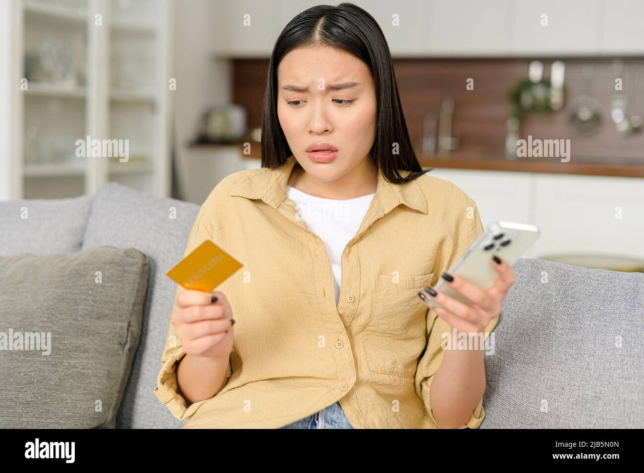 Frustrée femme asiatique insatisfaite assise sur un canapé, tenant le smartphone et le solde de carte de crédit. La jeune femme n'a pas assez d'argent pour les transactions en ligne, problème financier Banque D'Images