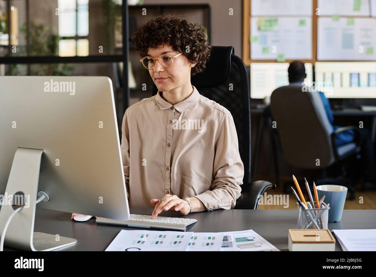 Jeune femme d'affaires en lunettes assise sur son lieu de travail devant un écran d'ordinateur et la dactylographie Banque D'Images