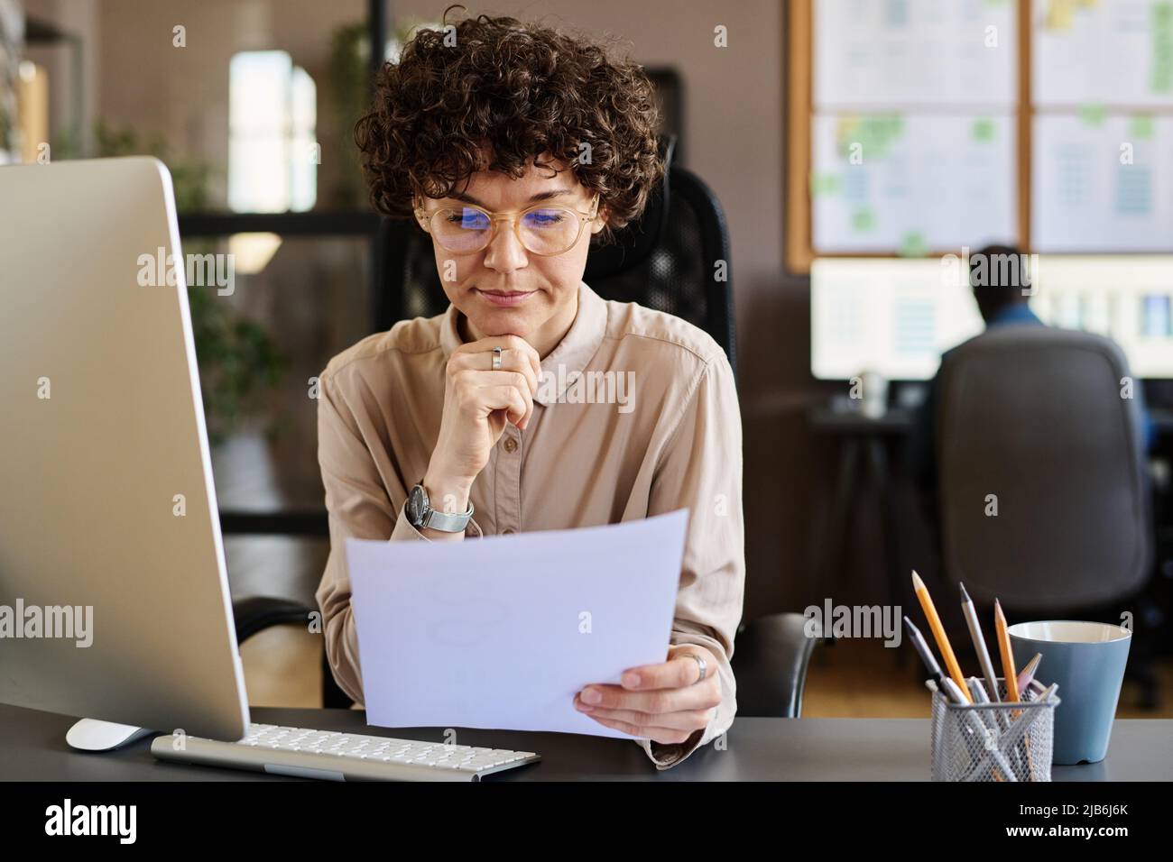 Jeune Manager en lunettes assis sur son lieu de travail devant l'ordinateur et le contrat de lecture avec une expression sérieuse Banque D'Images