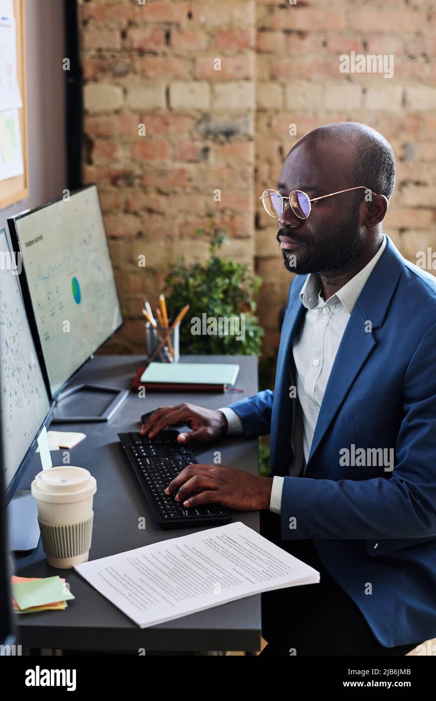 Responsable africain sérieux de lunettes assis sur son lieu de travail au bureau et dactylographiant, il travaille avec des cartes sur ordinateur Banque D'Images