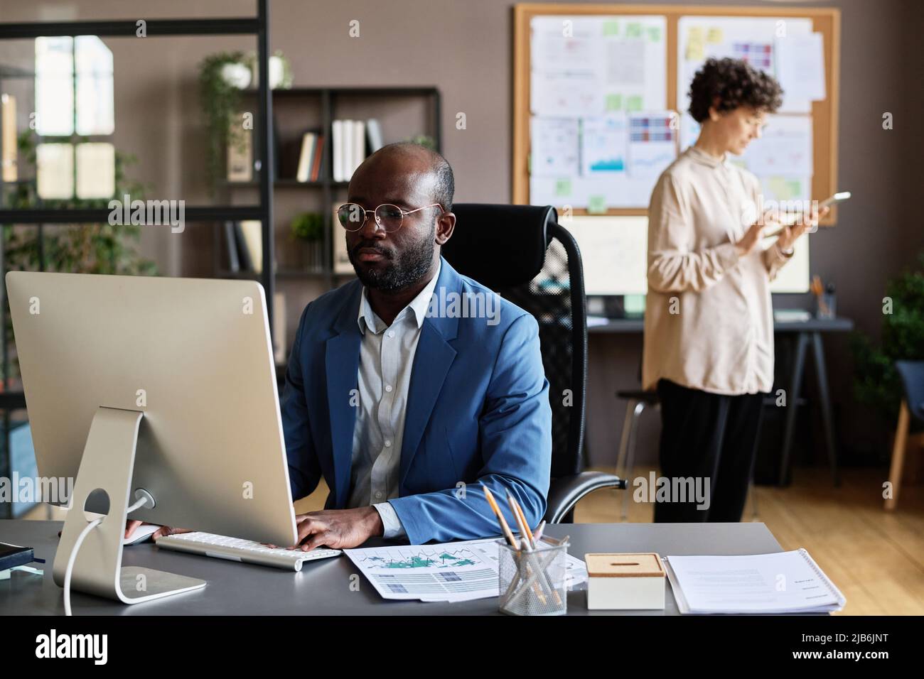 Manager africain assis sur son lieu de travail et se concentrant sur le travail en ligne sur l'ordinateur avec une femme debout en arrière-plan Banque D'Images