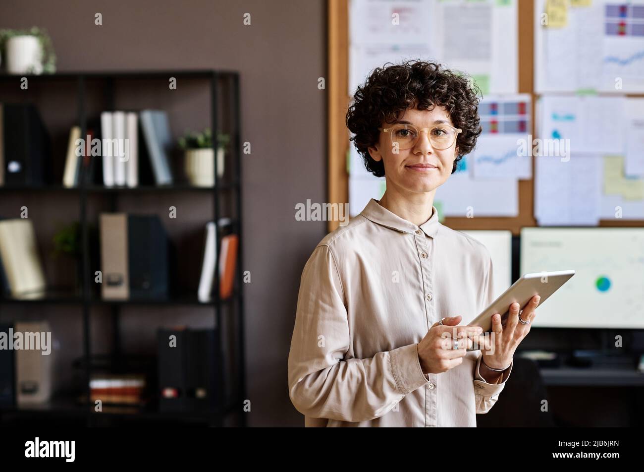 Portrait d'un jeune employé dans des lunettes regardant l'appareil photo tout en utilisant une tablette numérique au travail Banque D'Images