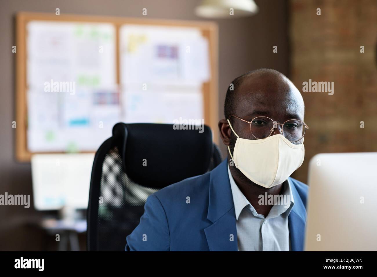 Homme d'affaires africain sérieux en lunettes et masque de visage travaillant sur ordinateur à son lieu de travail au bureau Banque D'Images