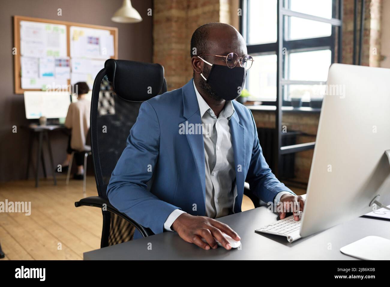 Un jeune Manager africain sous masque facial assis sur son lieu de travail devant un moniteur d'ordinateur et en tapant sur le clavier au bureau pendant une pandémie Banque D'Images
