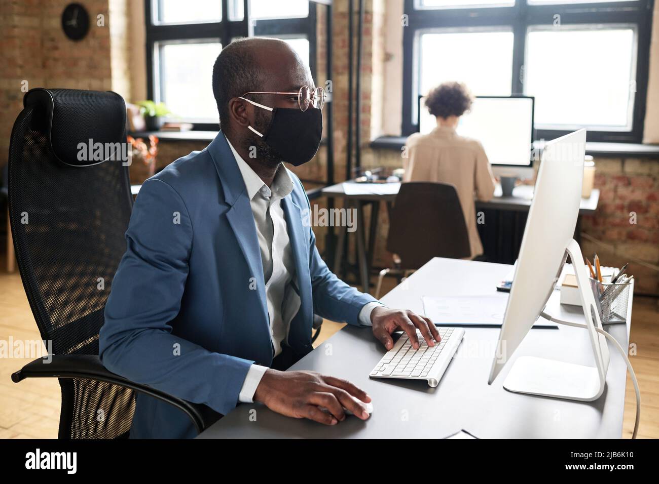 Homme d'affaires africain en costume et masque assis sur une chaise de bureau et travaillant sur un ordinateur à son lieu de travail au bureau Banque D'Images