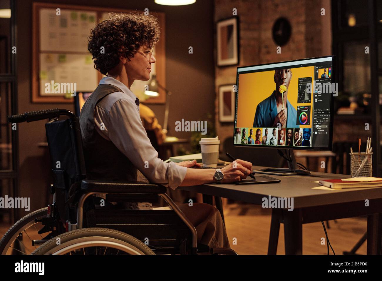 Jeune femme handicapée assise en fauteuil roulant sur son lieu de travail et travaillant au bureau comme graphiste Banque D'Images