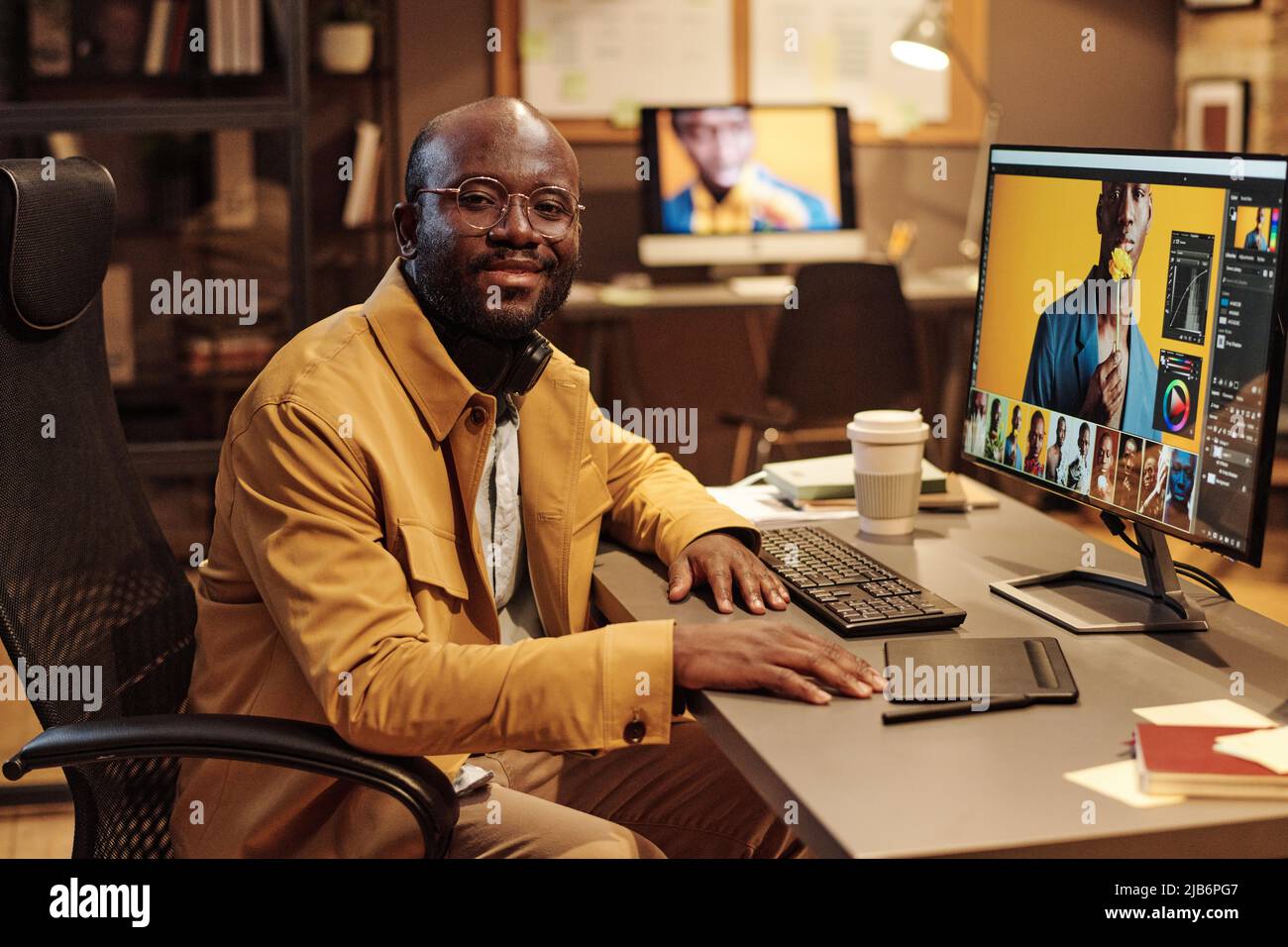 Portrait d'un graphiste africain en lunettes souriant à l'appareil photo tout en travaillant à table sur un ordinateur avec de nouveaux contenus Banque D'Images
