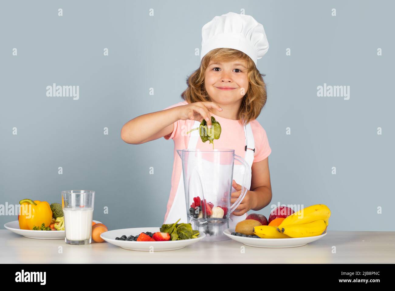 Portrait d'un enfant de 7, 8 ans en chapeau de cuisinier et tablier faire de la salade de fruits avec des épinards et cuisiner dans la cuisine. Joli petit blond joyeux sourire Banque D'Images