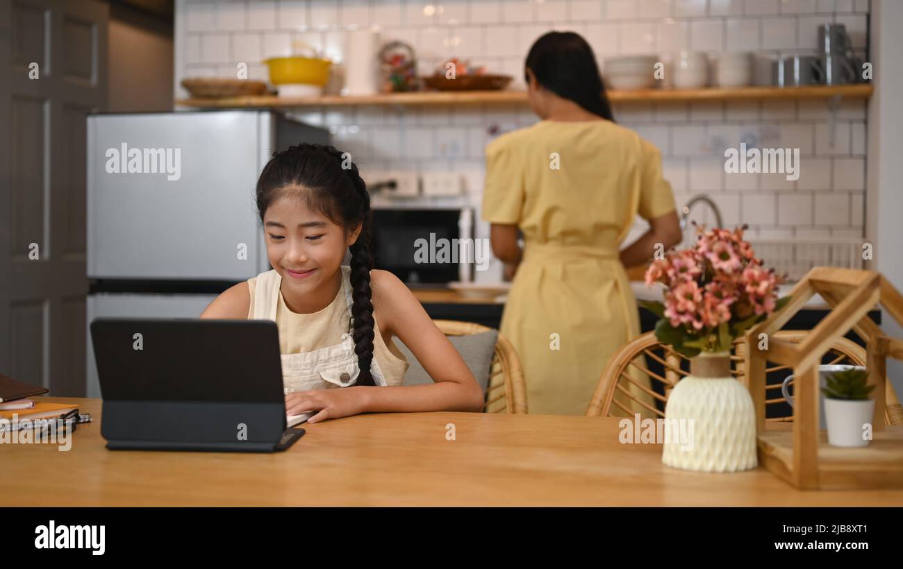 Fille asiatique ayant cours en ligne sur tablette d'ordinateur, faisant des devoirs à la table de cuisine. Concept d'éducation virtuelle, homeschooling Banque D'Images