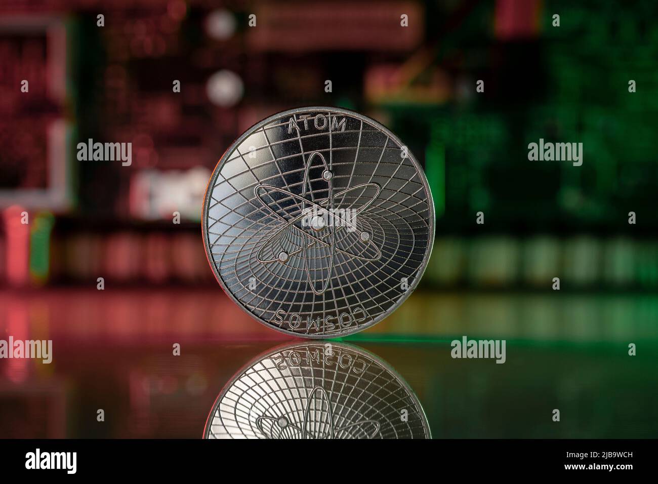 La pièce physique de crypto-monnaie ATOM COSMOS est placée sur une surface réfléchissante avec un micro-schéma en arrière-plan et est éclairée par des lumières vertes et rouges. Banque D'Images