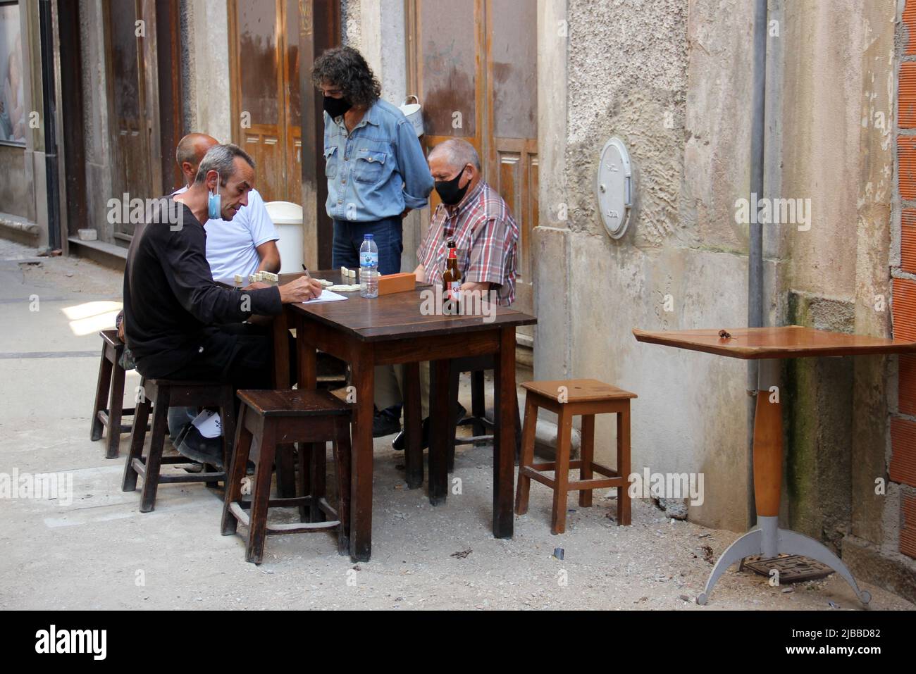 Quatre hommes, portant des masques, jouant au jeu de dominos à une table en bois dans la rue de la vieille ville, Coimbra, Portugal Banque D'Images