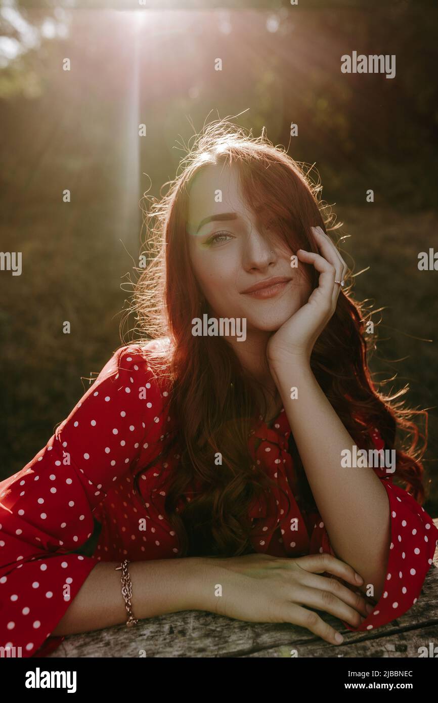 Gros plan visage portrait beauté blanc caucasien avec les cheveux longs rouges bouclés vêtus de chemise rouge tenant sa tête à la main et regardant l'appareil photo Banque D'Images