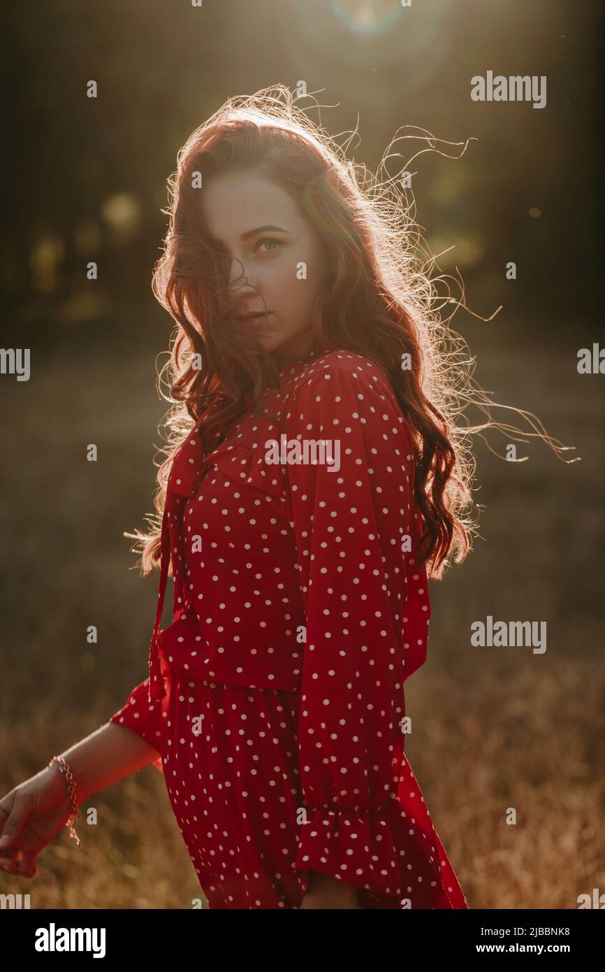 Portrait moyen de la jeune femme à tête rouge en robe rouge avec des points blancs sur fond de contre-jour de coucher de soleil. Modèle féminin regardant l'appareil photo Banque D'Images