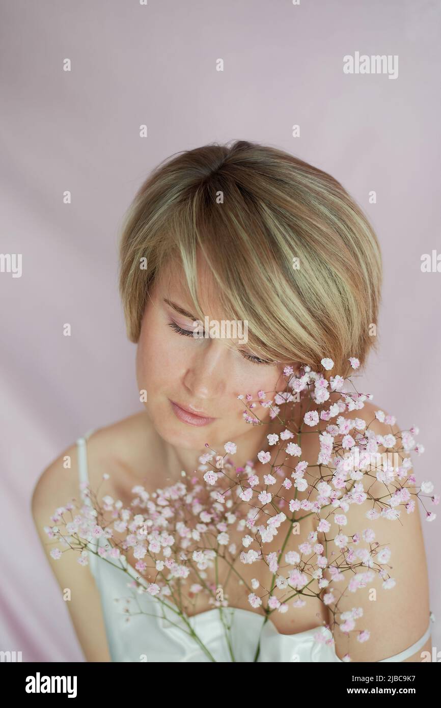 Portrait d'une fille sur fond rose avec des fleurs. Tendresse et féminité. La sphère de la beauté et la santé des femmes. Coupe courte, cheveux blonds Banque D'Images
