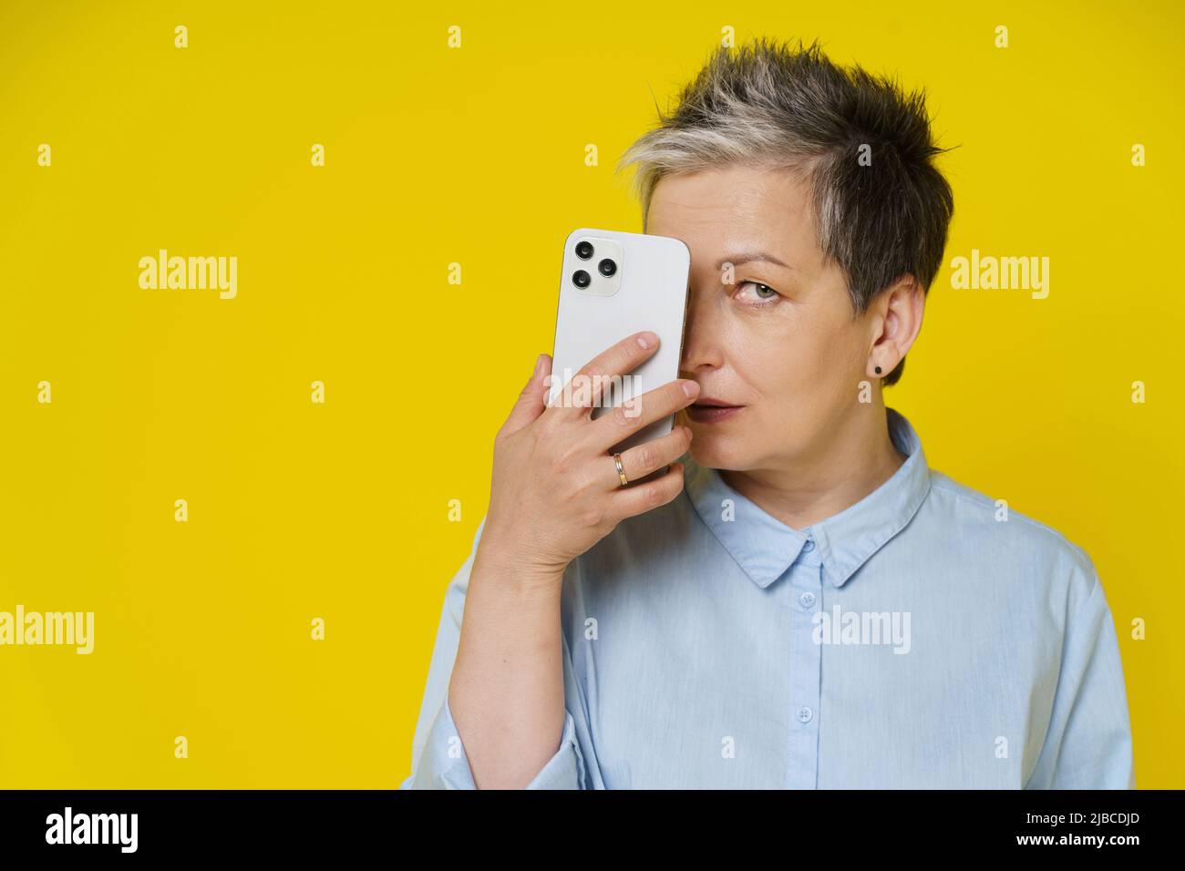 Une femme mûre grise cache le visage derrière un smartphone en publiant des commentaires négatifs et toxiques en ligne, sur les réseaux sociaux. Jolie femme avec téléphone en chemise bleue isolée sur fond jaune. Banque D'Images