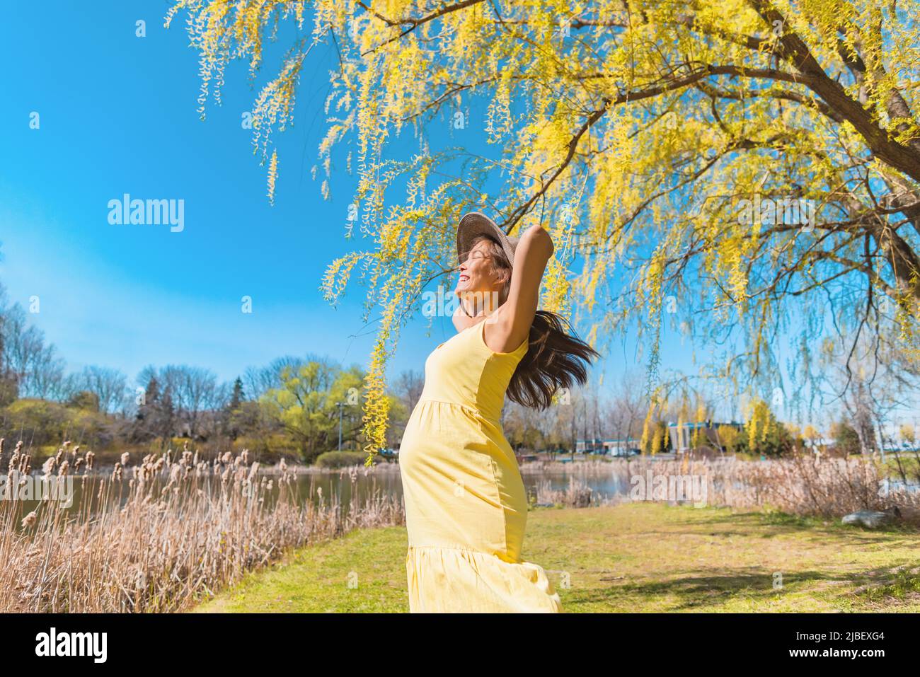 Bonne femme enceinte asiatique dansant gratuitement dans la nature parc d'été portant chapeau de soleil et robe jaune. Grossesse joie et insouciance Banque D'Images