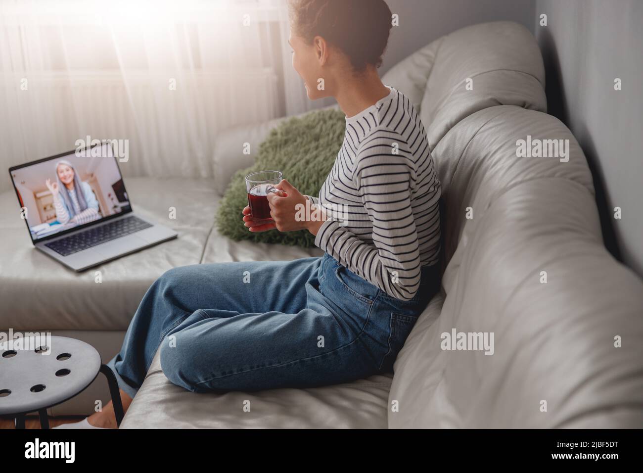 Une jeune femme est assise sur un canapé en cuir dans une chambre confortable avec une tasse de thé et un appel vidéo sur un ordinateur portable Banque D'Images