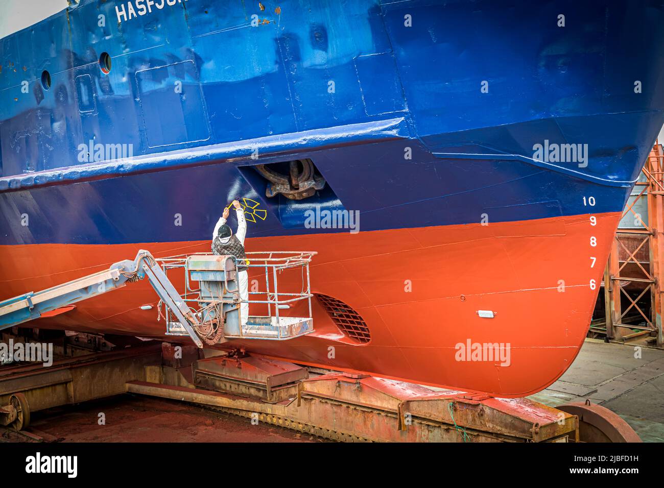 Un travailleur en combinaison de protection, debout sur une plateforme élévatrice mobile, repeint la marque jaune de propulseur d'étrave sur la coque bleu foncé et rouge du ferry roulier Hasfjord en cale sèche à l'usine MEST de Tórshavn, îles Féroé. Banque D'Images