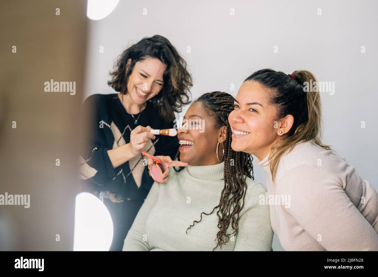 Des amies multiraciales en vêtements décontractés souriant et regardant le miroir pendant la séance de maquillage dans le salon de beauté Banque D'Images