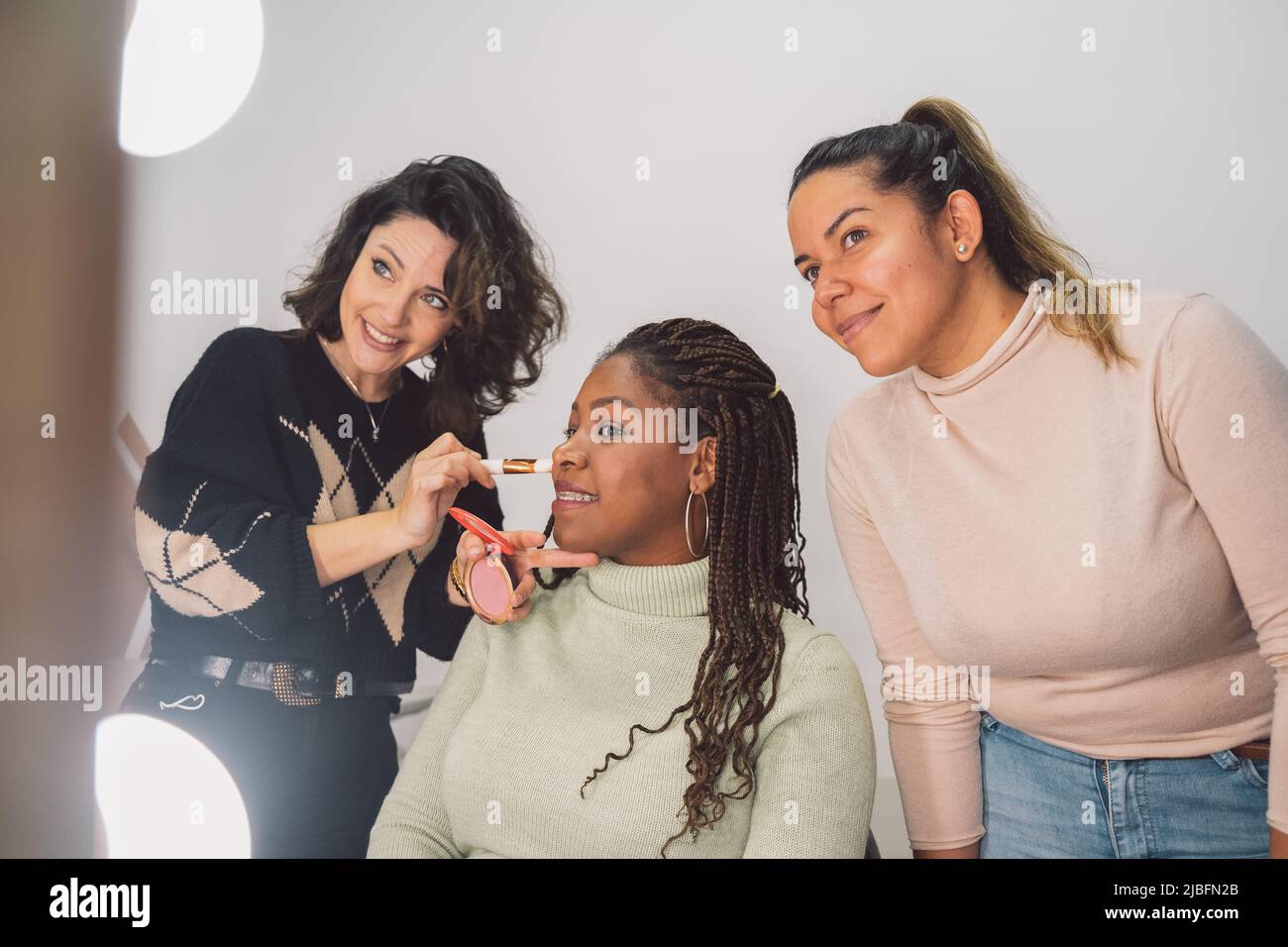 Des amies multiraciales en vêtements décontractés souriant et regardant le miroir pendant la séance de maquillage dans le salon de beauté Banque D'Images