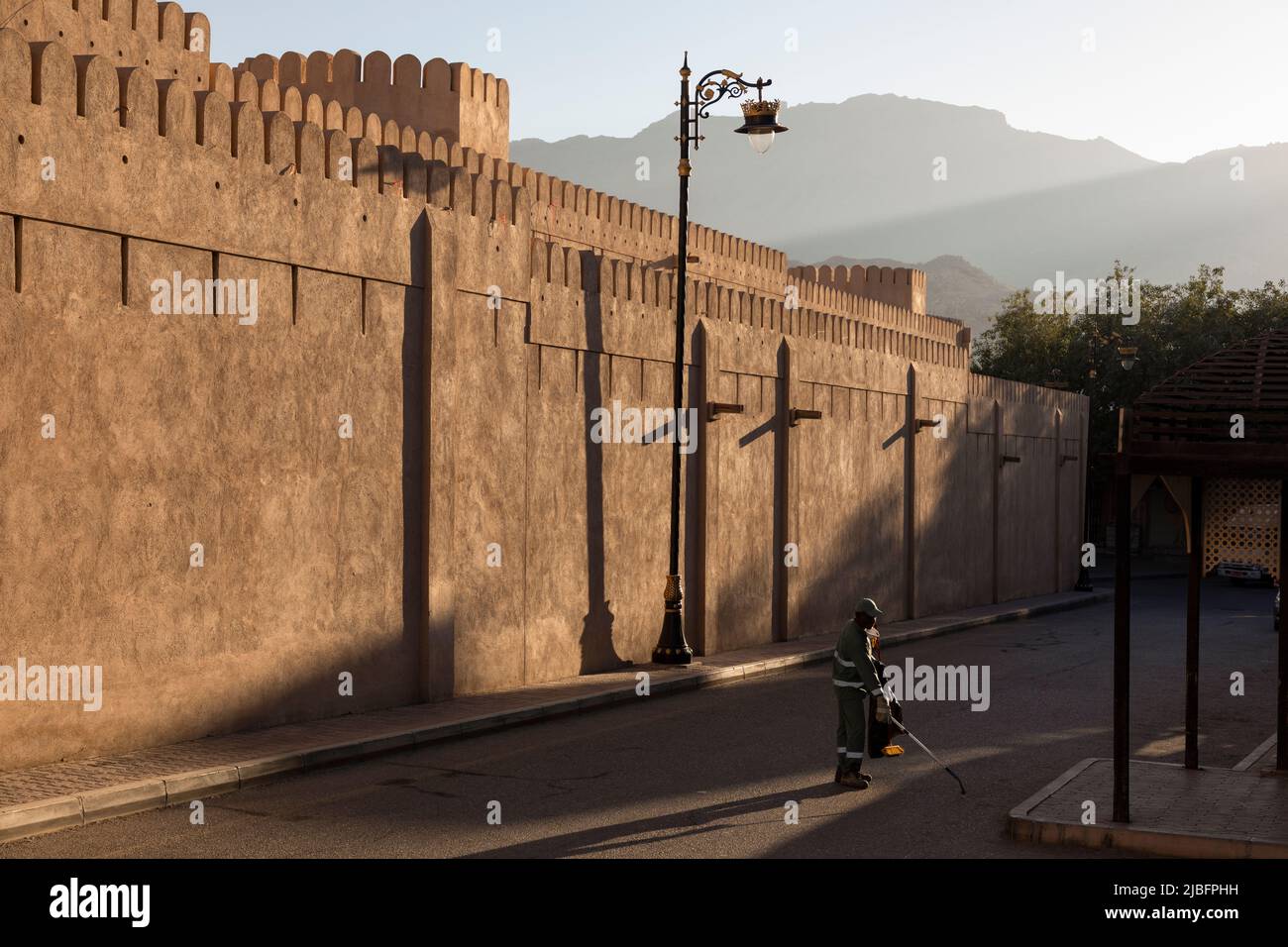Un nettoyeur de rue travaillant à côté des murs historiques de Nizwa, Oman, dans la lumière tôt le matin avec de longues ombres et une architecture traditionnelle. Banque D'Images
