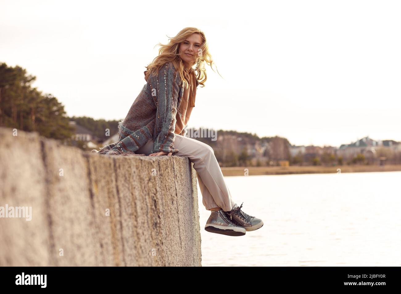 Portrait d'une jeune femme souriante avec de longs cheveux ondulés et justes assis avec des jambes croisées sur le bord de la rive de rivière en béton. Banque D'Images