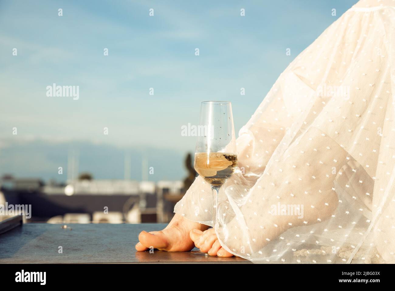 Photo courte des jambes pieds nus d'une femme vêtue d'une longue robe d'été blanche et transparente, avec un verre de champagne. Banque D'Images