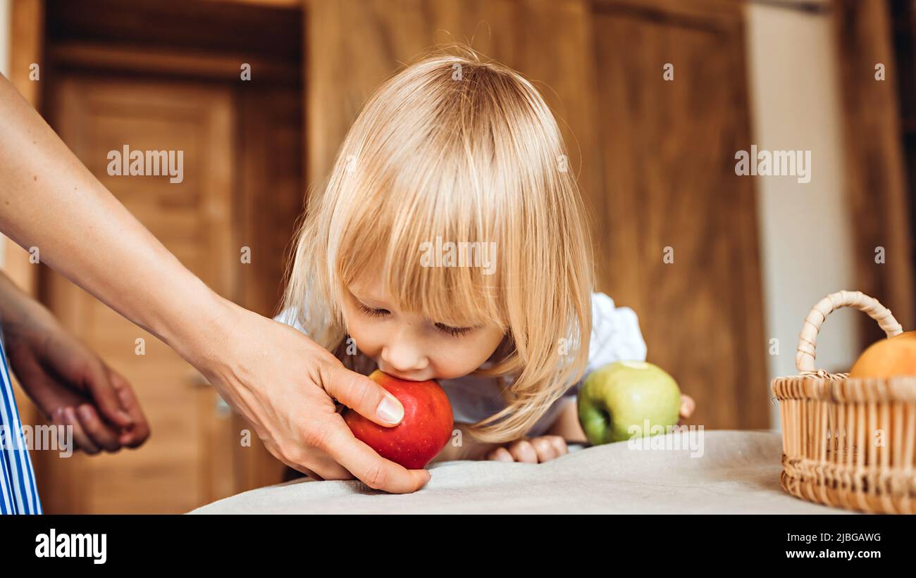 Une petite fille mord une pomme qui est tenue par une maman ou une gardienne assise à la table Banque D'Images