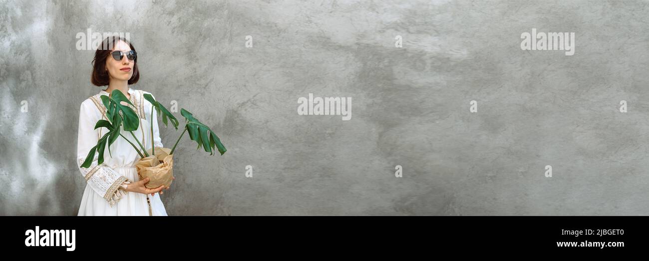 Baner fait avec photo la fille dans le beau blanc style boho tenue en pot plante Monstera avec de charmantes feuilles vertes sur un fond de mur en béton à la mode. Femme en lunettes de soleil. Copier l'espace Banque D'Images