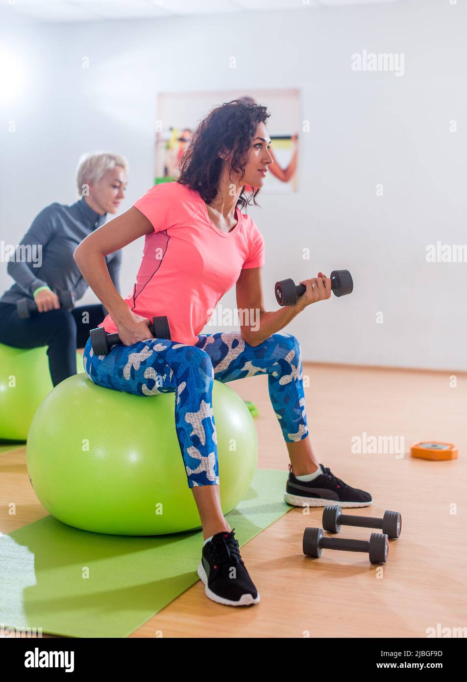 Sportives femmes minces prenant part à la salle de gym classe de fitness s'exerçant assis sur des physiobales faisant alternés biceps curl avec des haltères. Banque D'Images