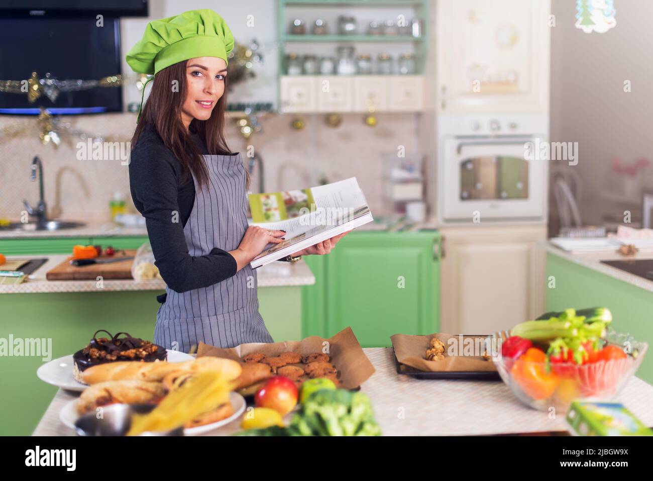 Jeune femme de ménage essayant de trouver une nouvelle recette dans le livre de cuisine tout en se tenant à table avec la nourriture et les ingrédients. Banque D'Images