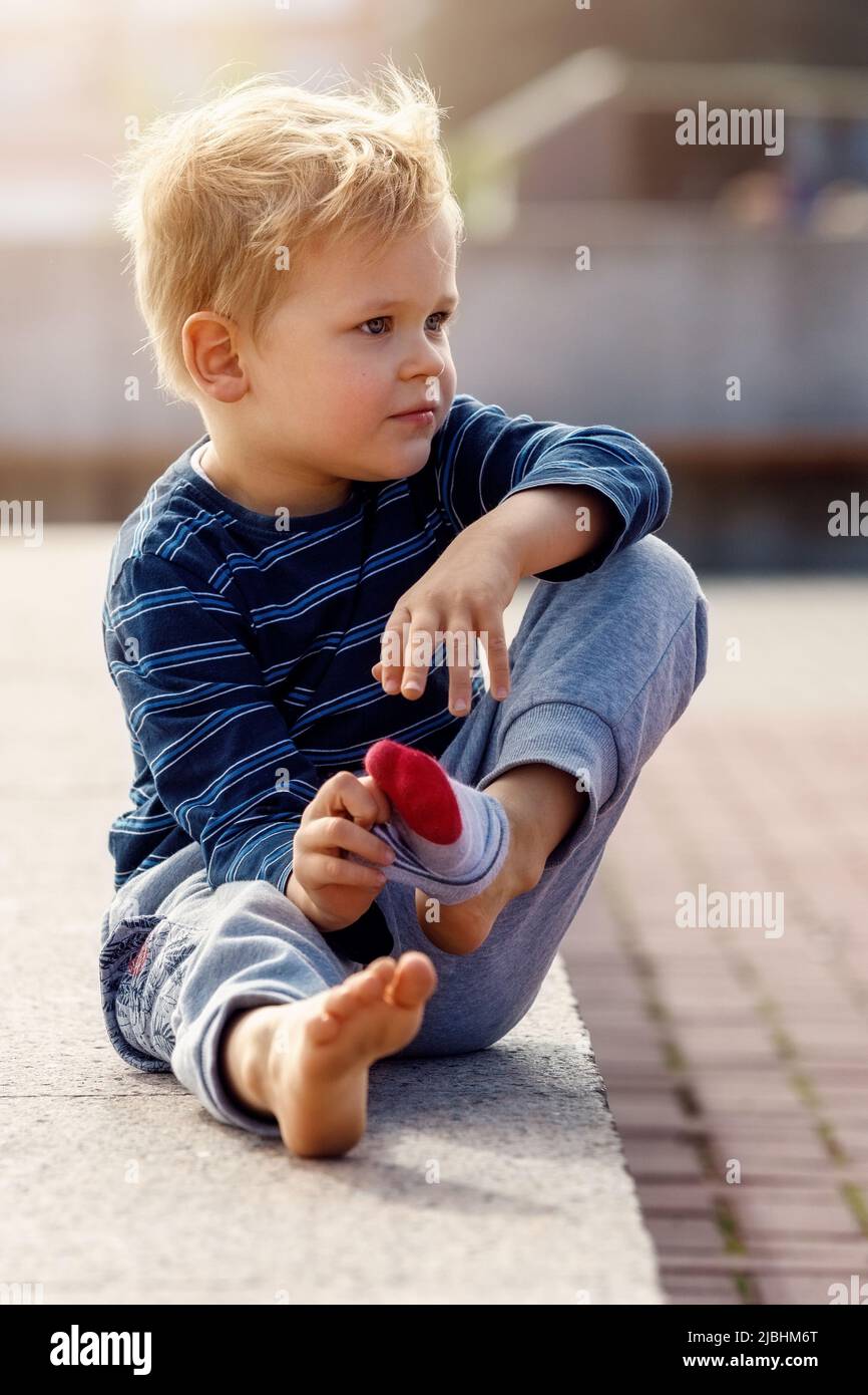 Un portrait d'un petit garçon mignon assis près d'une fontaine de la ville, l'enfant prend ses chaussettes, il veut refroidir ses pieds dans l'eau fraîche. Banque D'Images