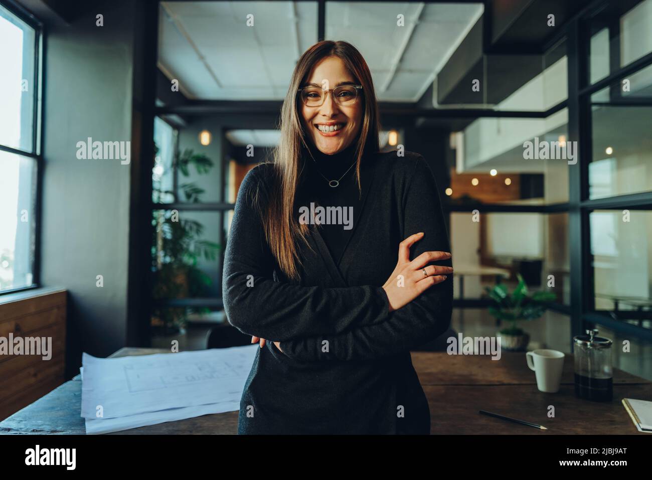 Une femme entrepreneure gaie souriant à la caméra tout en se tenant debout avec ses bras croisés. Bonne jeune femme d'affaires debout dans la salle de réunion d'un mode Banque D'Images