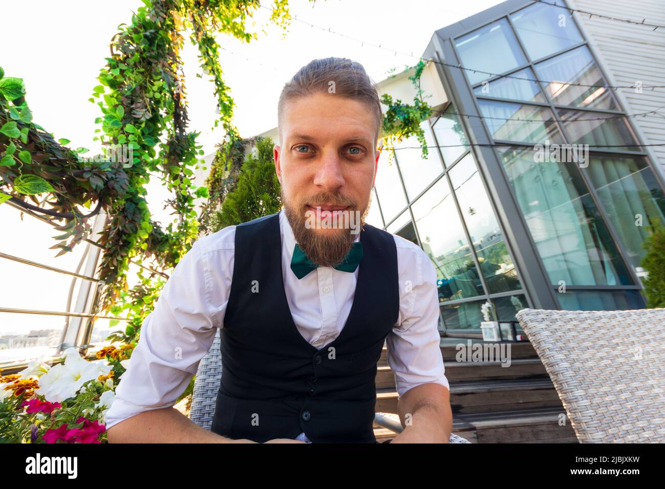 Jeune homme barbu dans une chemise blanche, gilet noir et noeud papillon regarde la caméra tout en étant assis dans un penthouse Banque D'Images