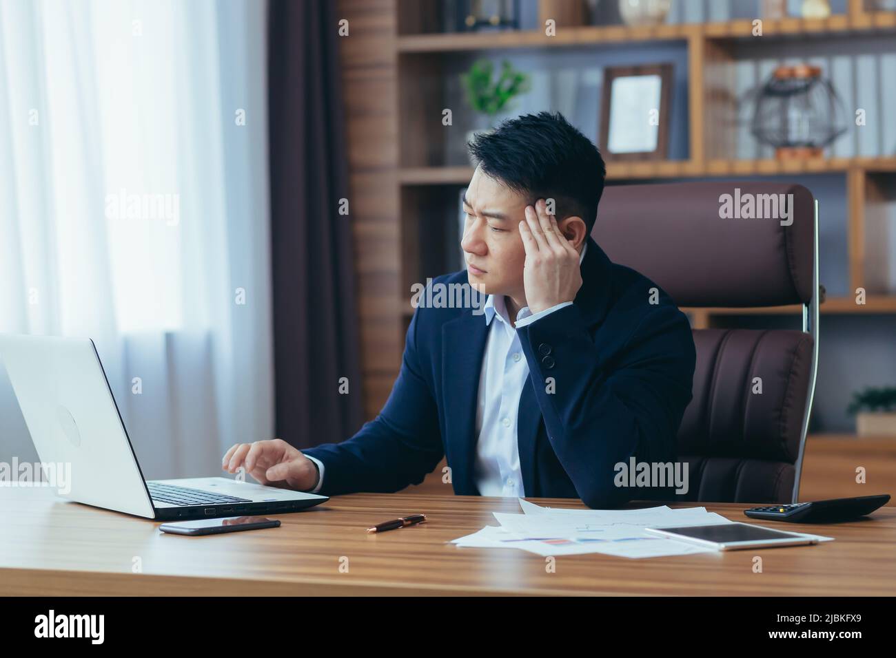 Jeune homme d'affaires asiatique fatigué, travailleur, avocat au travail. Il a un mal de tête, garde ses mains sur sa tête, s'assoit à un bureau dans le bureau Banque D'Images