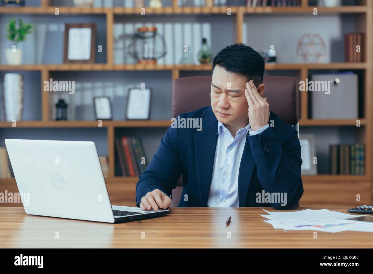 Jeune homme d'affaires asiatique fatigué, travailleur, avocat au travail. Il a un mal de tête, garde ses mains sur sa tête, s'assoit à un bureau dans le bureau Banque D'Images