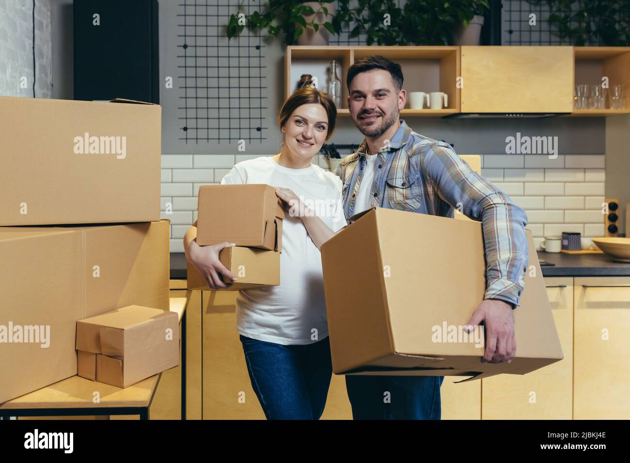 Portrait de jeune mari de famille et femme enceinte dans un nouvel appartement tenant des boîtes en carton regardant l'appareil photo et souriant Banque D'Images