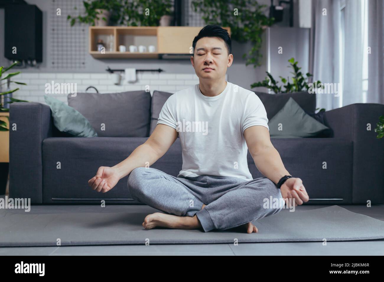 Asiatique lui-même médite à la maison le matin, assis sur un tapis de sport, près du canapé dans le salon, son matin, calme à l'extérieur du lotus Banque D'Images