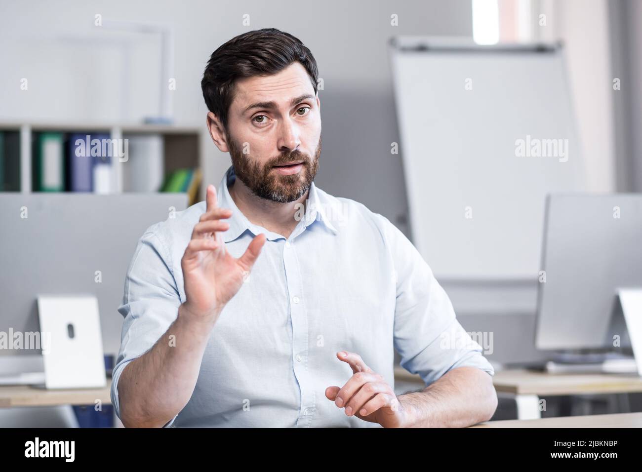 Jeune beau coach d'affaires réussi explique et dit sur la caméra, les gestes avec ses mains. Assis à un bureau d'ordinateur dans le bureau, regardant t Banque D'Images