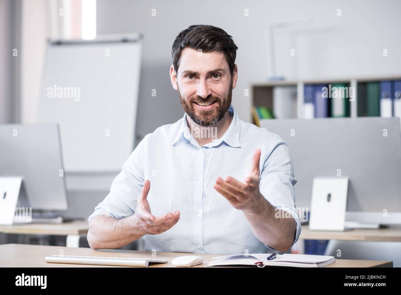 Jeune beau coach d'affaires réussi explique et dit sur la caméra, les gestes avec ses mains. Assis à un bureau d'ordinateur dans le bureau, regardant t Banque D'Images