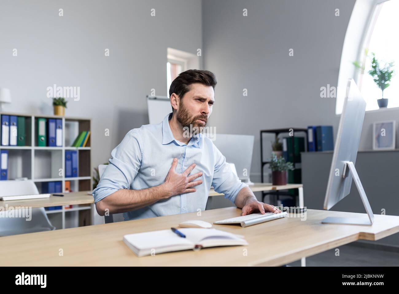 Un jeune homme fatigué travaillant à un ordinateur et assis à un bureau dans le bureau, ressent des douleurs à la poitrine, un essoufflement, de la difficulté à respirer Banque D'Images