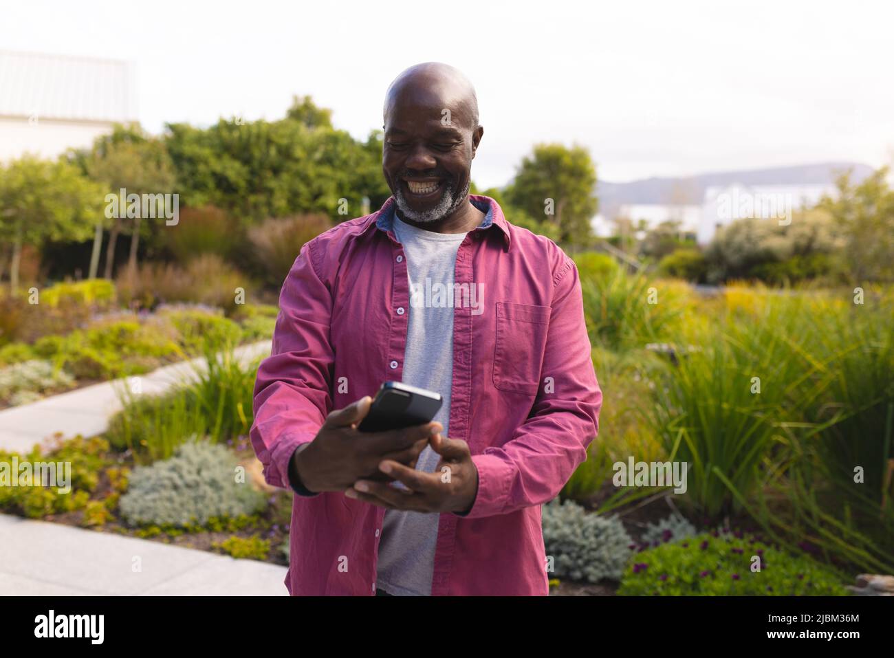 Homme âgé américain d'afrique chauve utilisant un téléphone portable tout en se tenant contre des plantes et ciel clair Banque D'Images