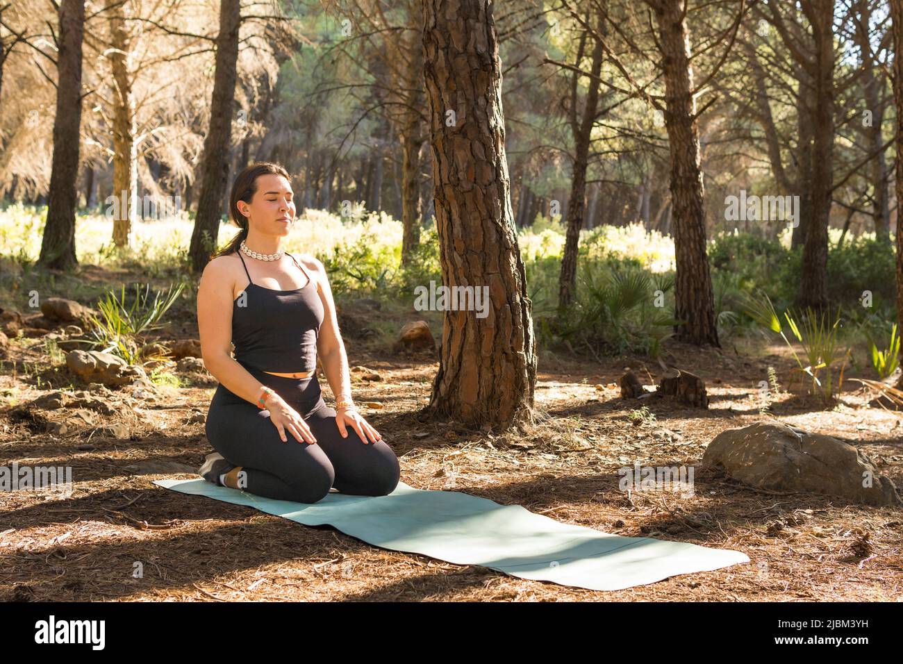 Belle jeune fille adulte faisant la méditation sur son tapis au coucher du soleil dans une forêt entourée d'arbres. Tir ouvert Banque D'Images